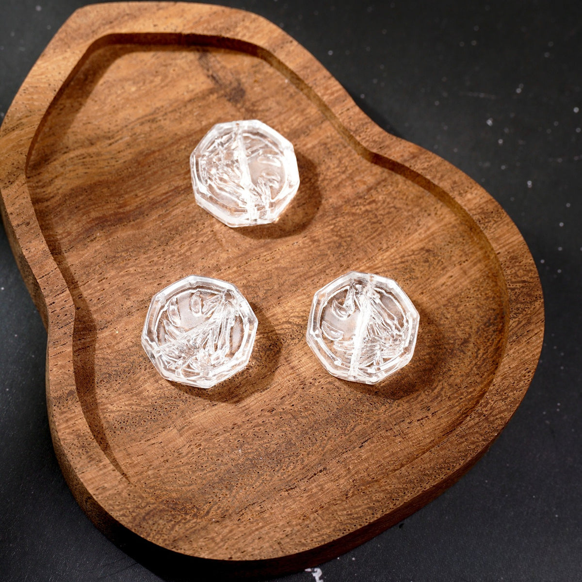 Three clear faceted engraved glass beads on a carved wooden tray against a dark background