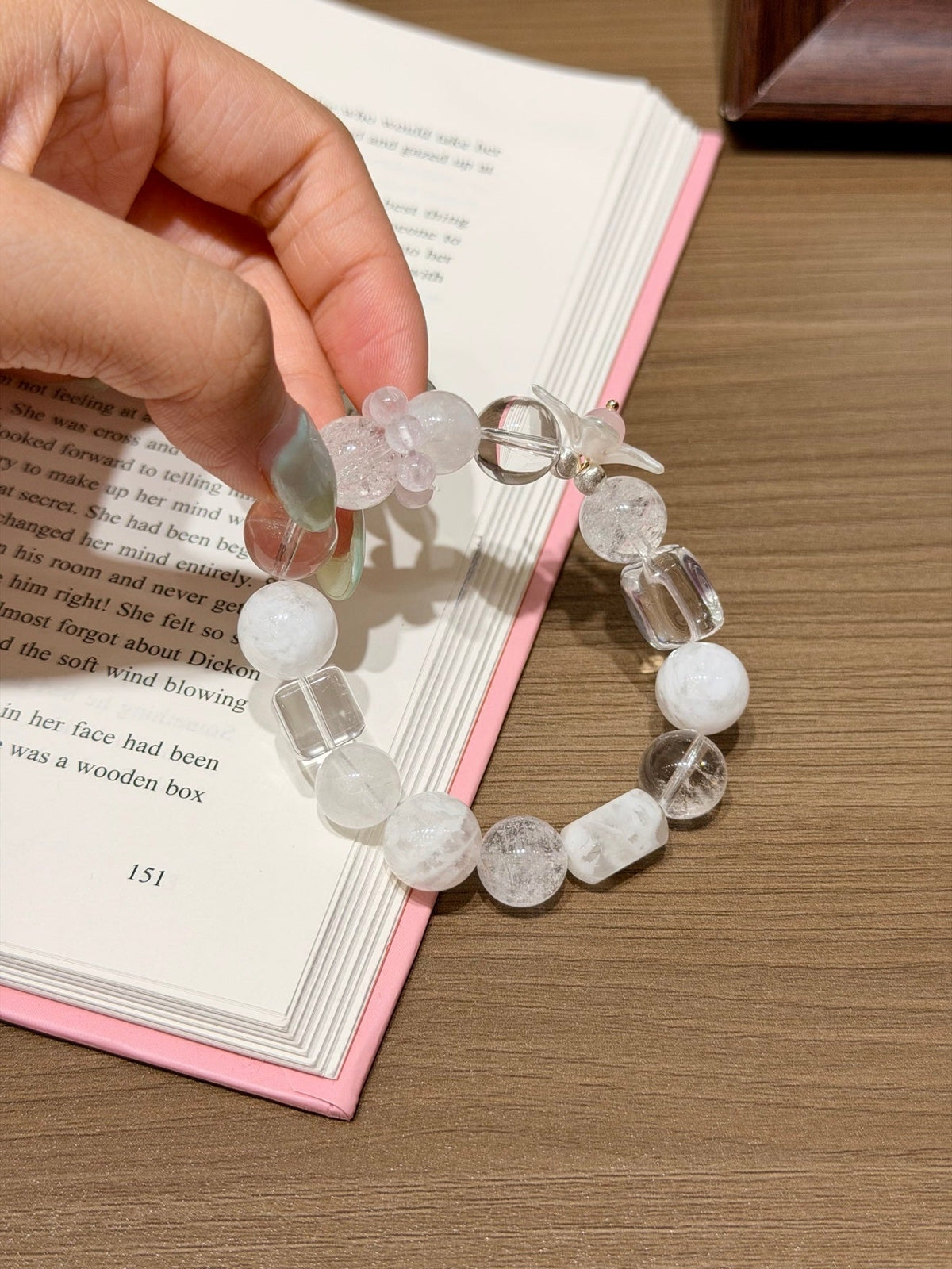 Hand holding a clear and white beaded bracelet with a mother-of-pearl flower charm over an open book