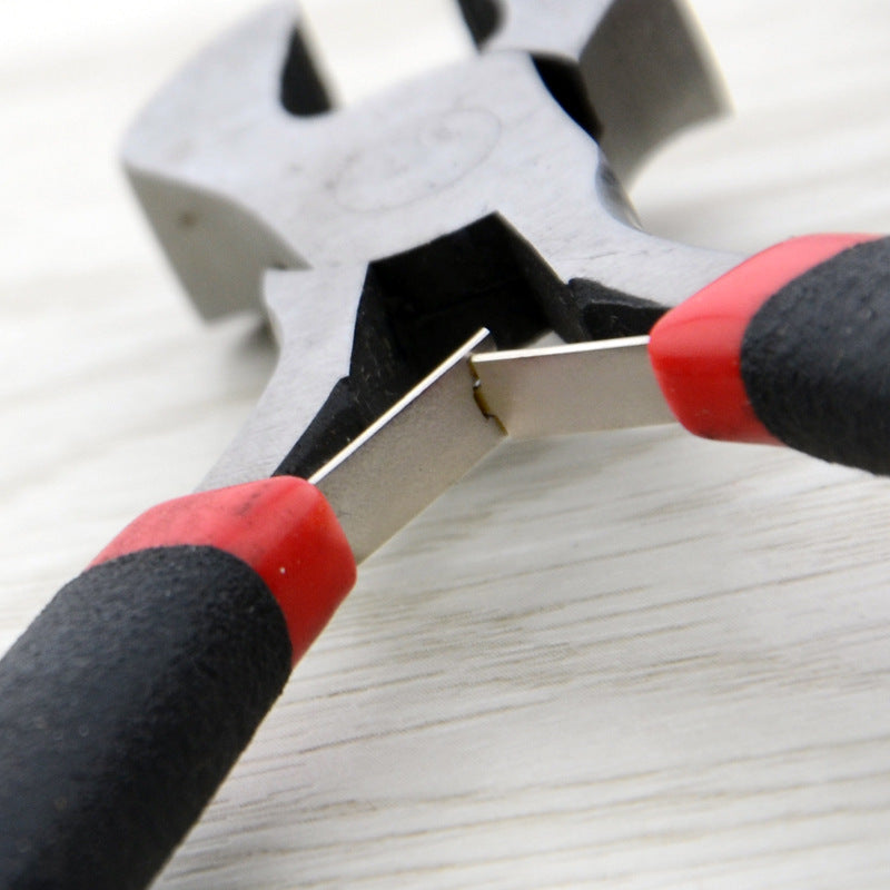 Close-up of a metal wire cutter or nippers with black and red handles, shown in action on a light wood surface.