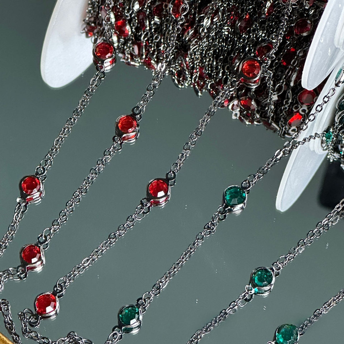 Silver-toned chain garlands draped over a reflective surface, decorated with alternating round red and green rhinestone beads in silver bezel settings.