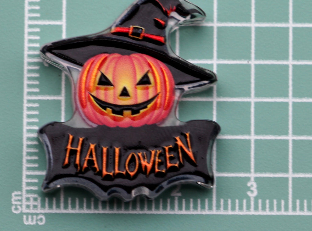 Halloween acrylic charm: smiling jack-o'-lantern in black witch hat above orange 'HALLOWEEN' banner on green cutting mat