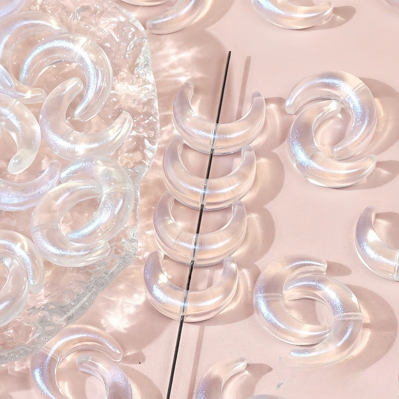 Clear iridescent glazed glass crescent moon beads, one strung on a black string and others scattered on a pink surface with a clear tray