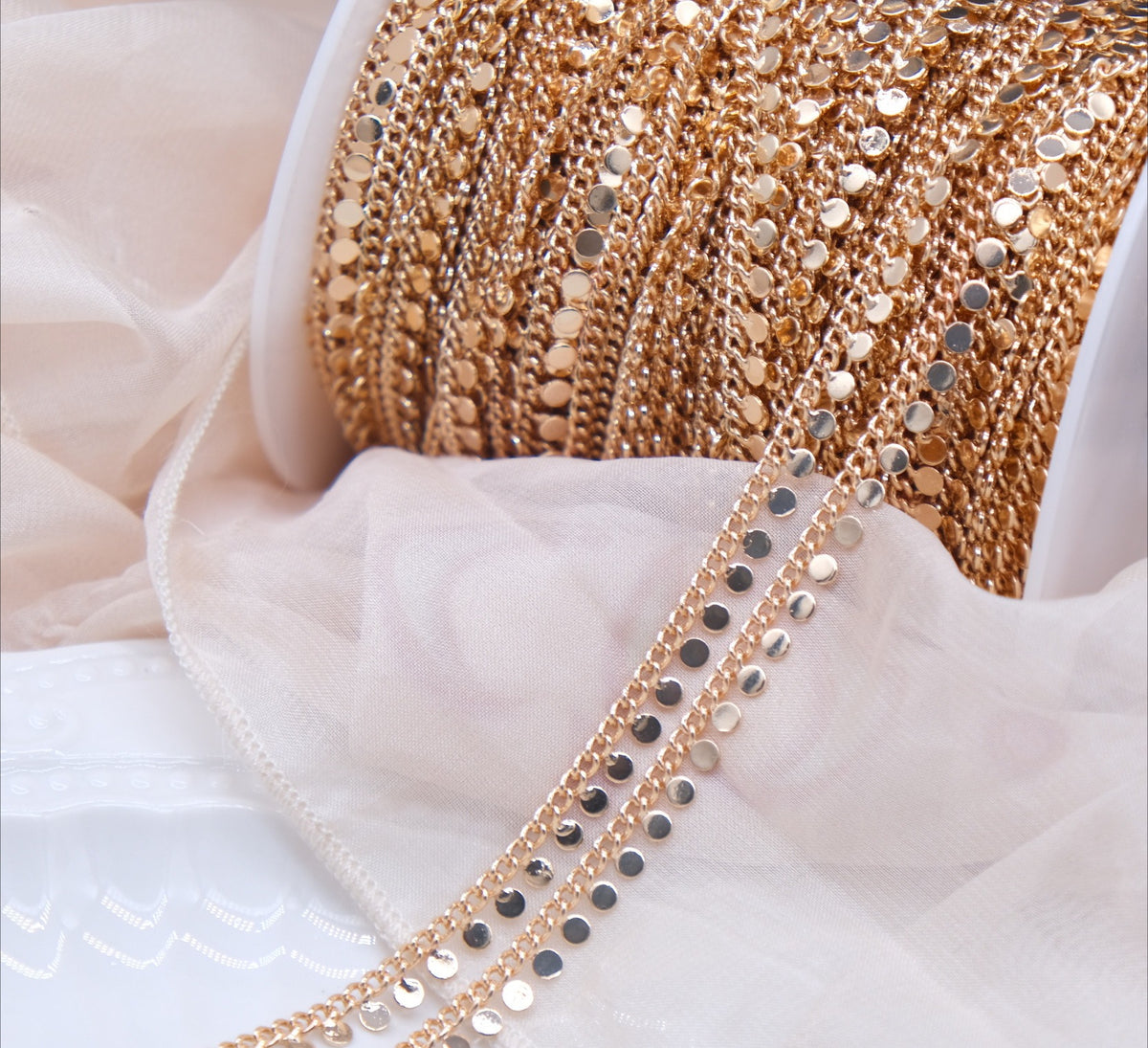 Spool of gold-plated disc chain with dangling round discs draped over white fabric.
