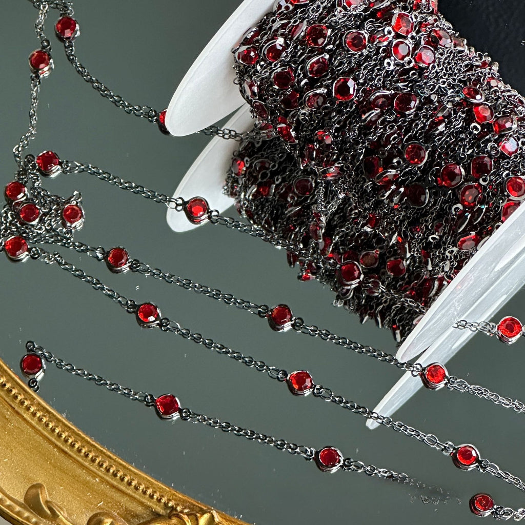 Spool of dark metal chain with red rhinestones, strands draped over a mirror.