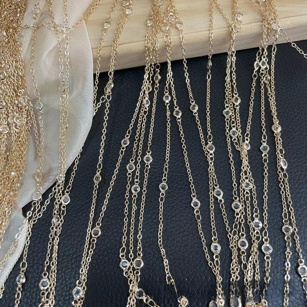 Gold-toned chain fringe garments with clear rhinestone or crystal bead accents, draped over a light wood surface and a dark textured background.