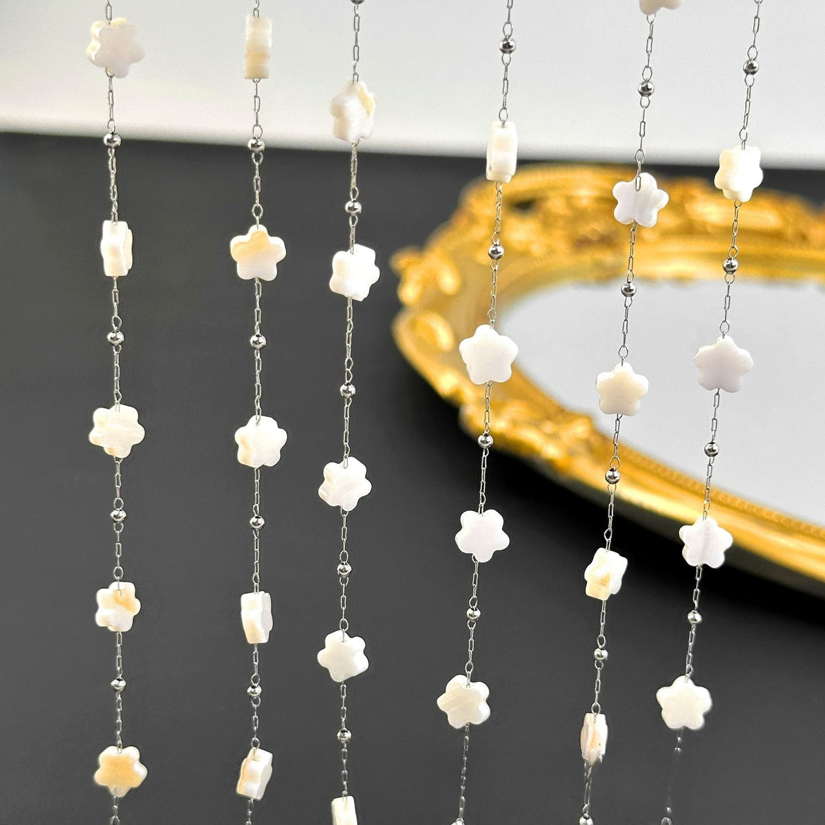 Multiple strands of white flower-bead station chain with small silver beads hanging before a dark surface.