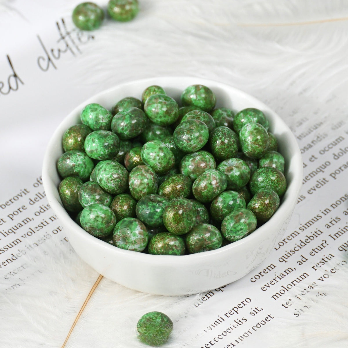 Green marbled round beads piled in a small white bowl on printed paper, a few loose beads nearby
