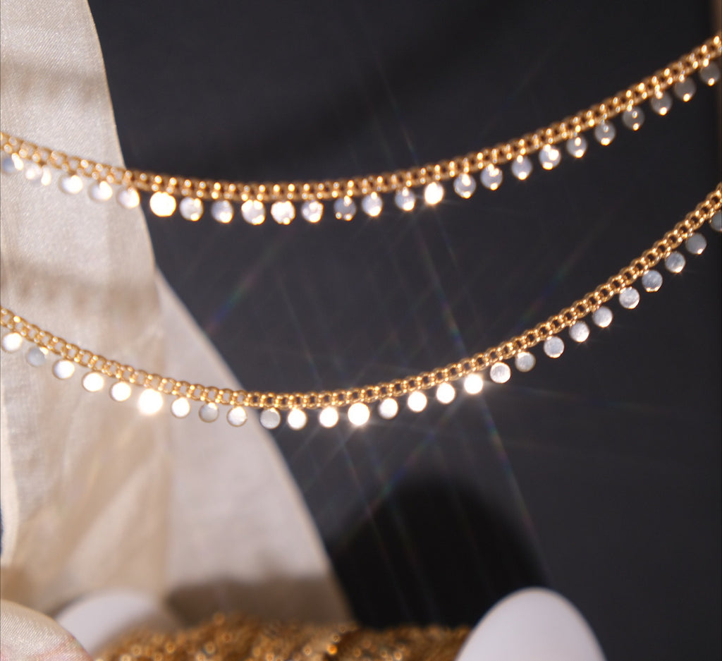 Gold disc chain with dangling round discs draped over a dark background beside beige fabric.