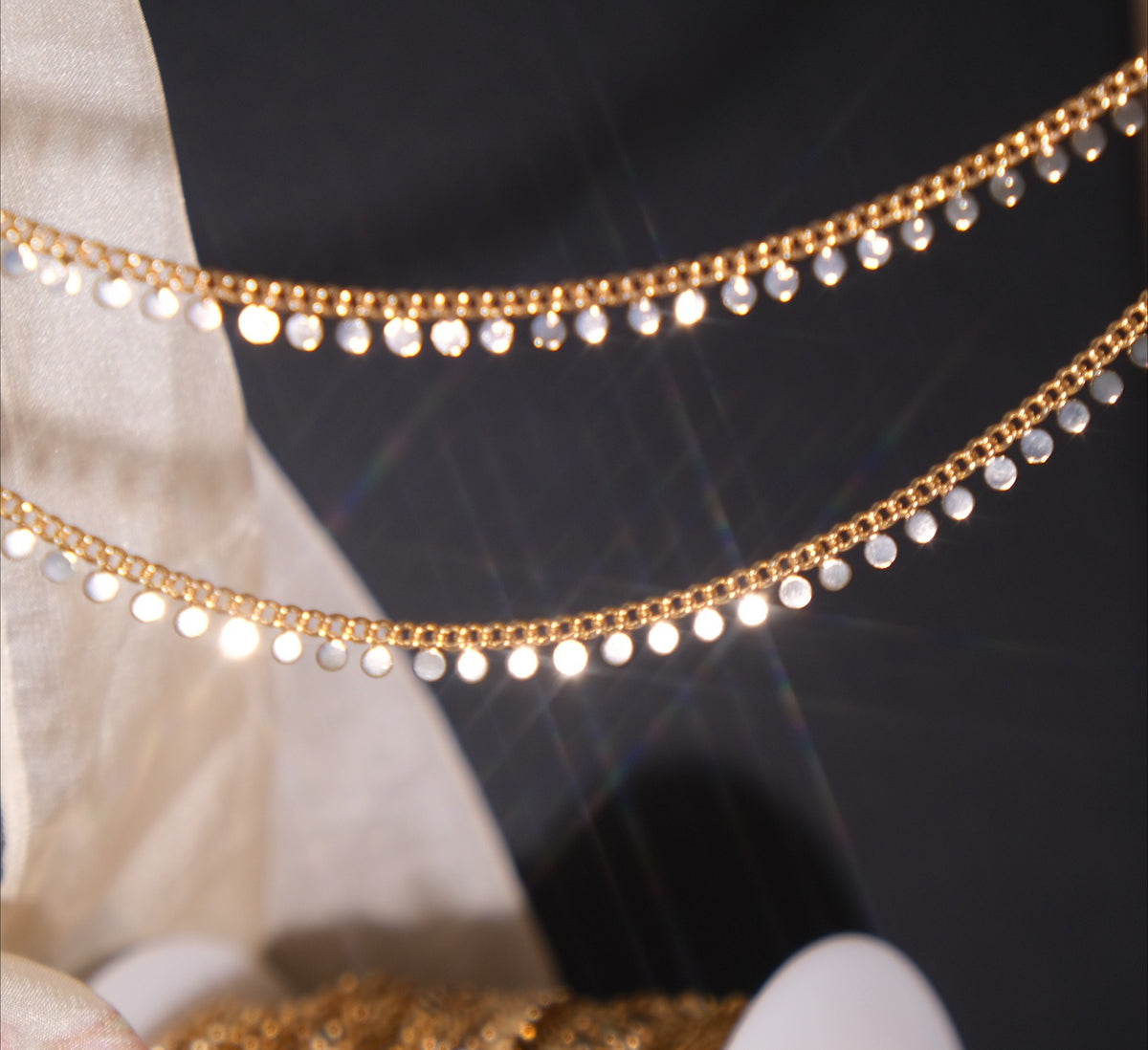 Gold disc chain with dangling round discs draped over a dark background beside beige fabric.