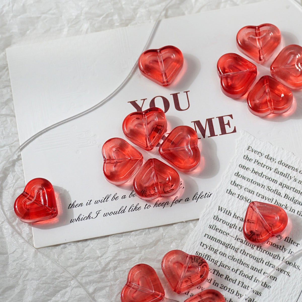 Red translucent heart-shaped beads scattered on white printed cards over a textured white fabric background.
