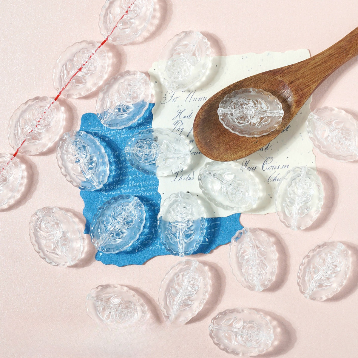 Clear oval glass beads with embossed floral details on pink with a wooden spoon and blue paper