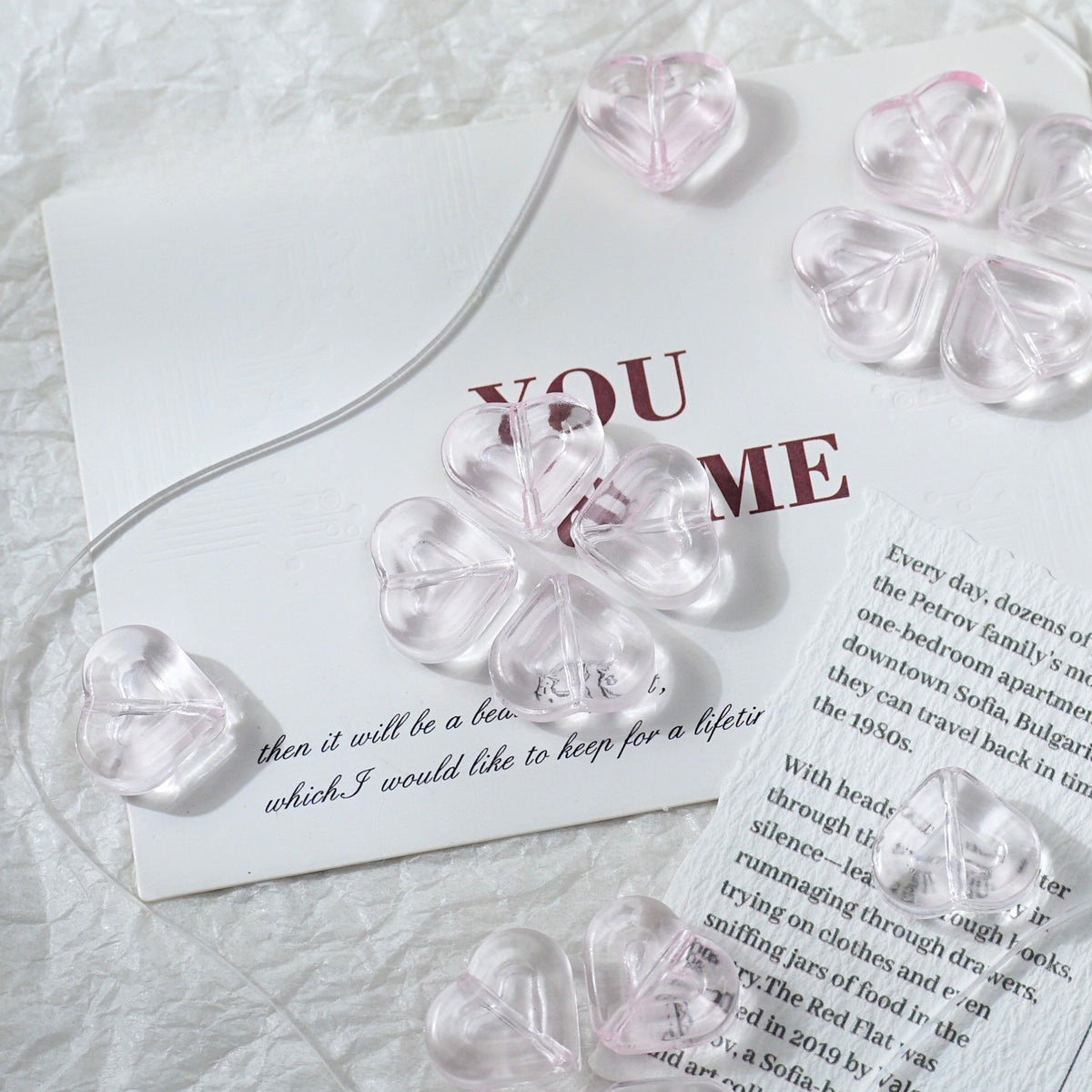Clear heart-shaped glass beads threaded on a transparent string, arranged on white textured paper with printed text.
