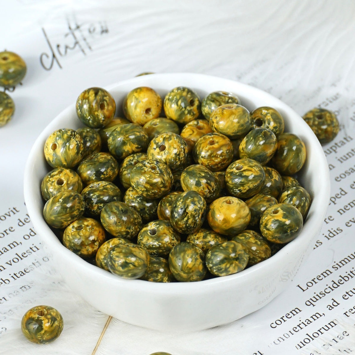 White porcelain bowl of marbled olive-green and yellow round beads on printed paper, a few loose beads scattered around
