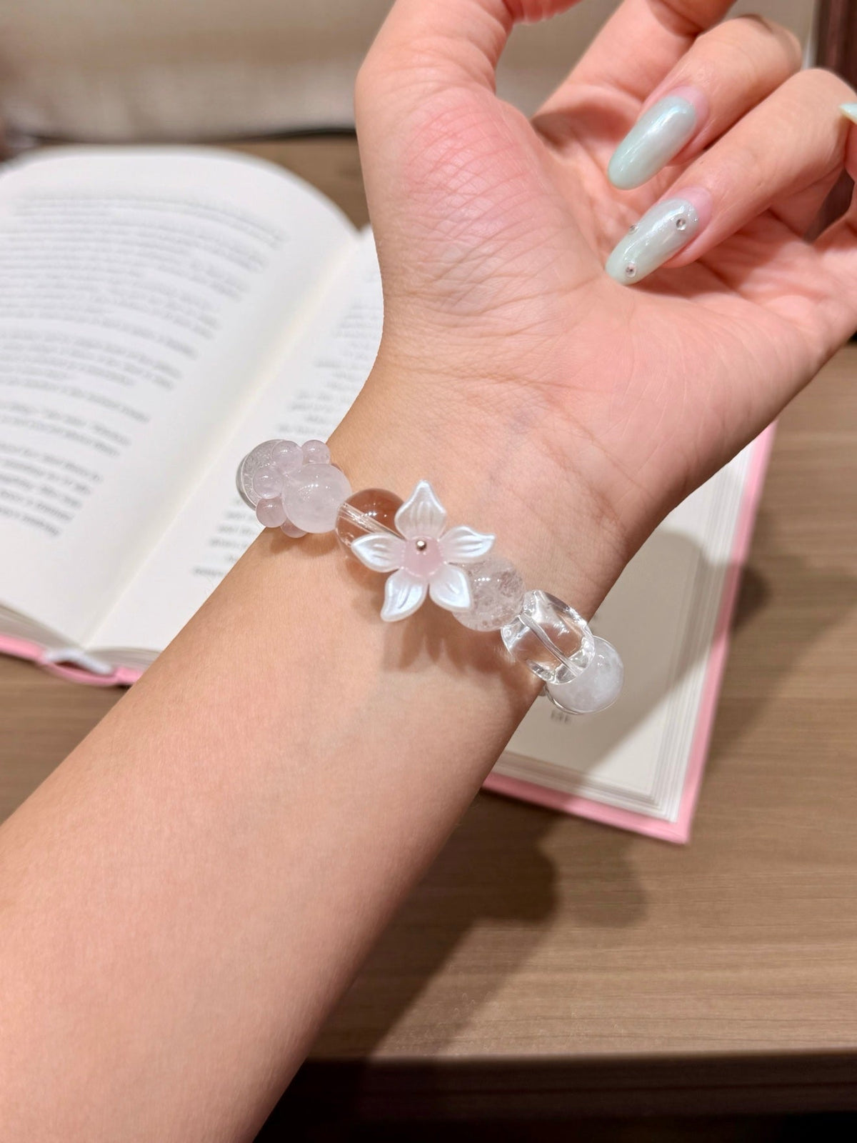 Translucent pink and white beaded bracelet with white flower charm worn on a wrist, open book behind