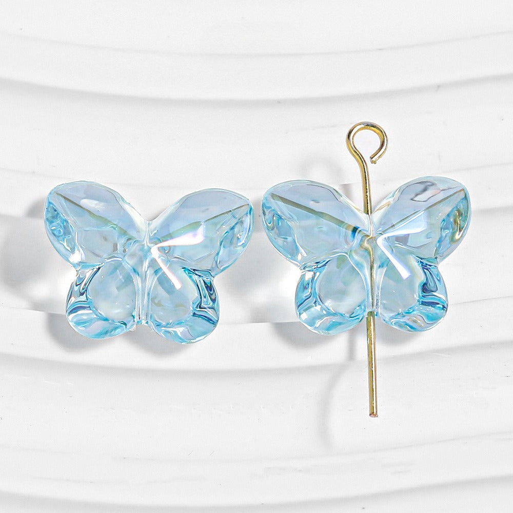 Two light blue faceted butterfly-shaped beads, one threaded on a gold headpin, on a white textured background