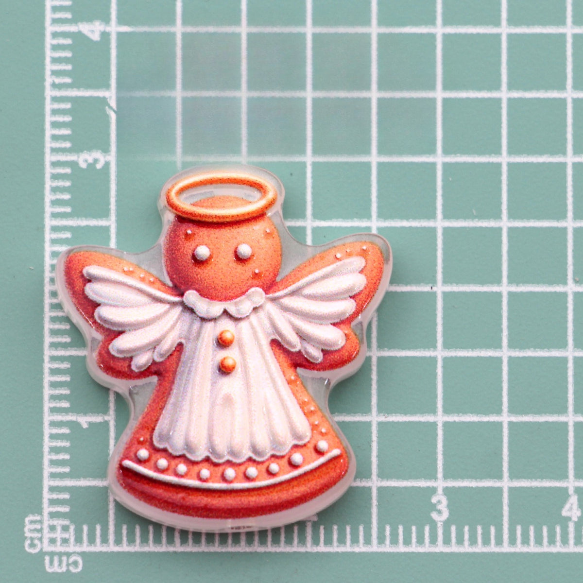 Gingerbread angel acrylic charm with white wings, dress and gold halo on green cutting mat