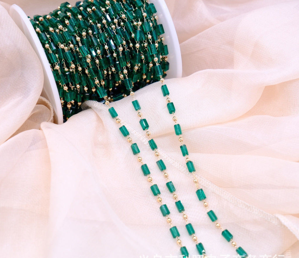 A spool of green faceted bead strands with small gold spacer beads, draped over soft cream-colored fabric.
