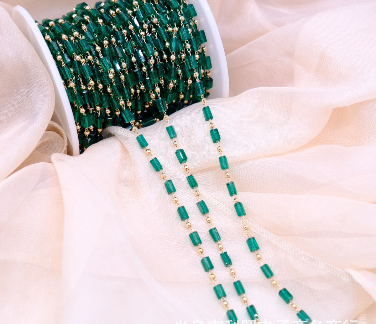 A spool of green faceted bead strands with small gold spacer beads, draped over soft cream-colored fabric.