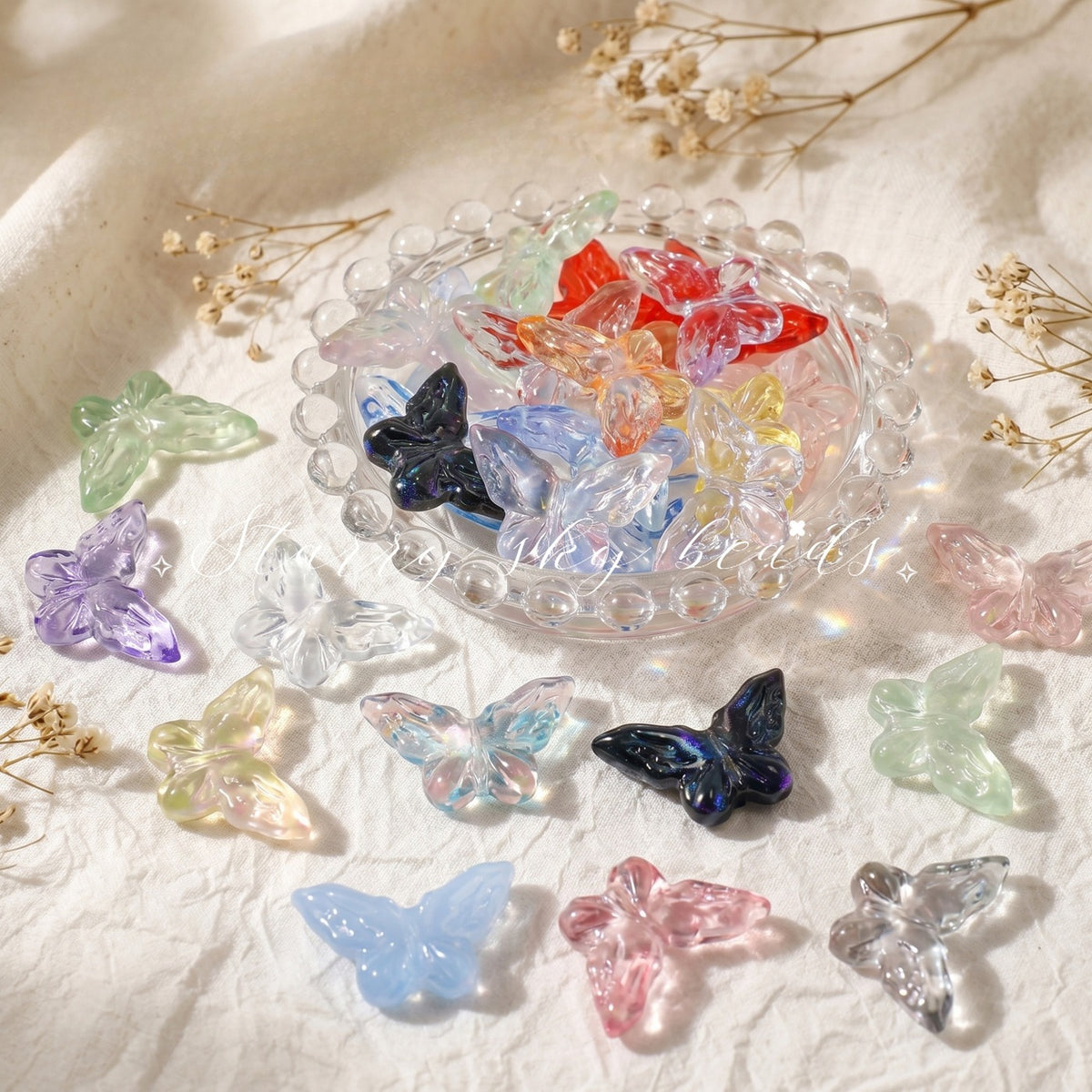 Translucent butterfly-shaped glass beads in assorted colors arranged on white fabric with dried flowers