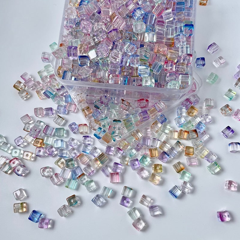 Pastel iridescent cube glass beads in a clear container, spilled on white surface