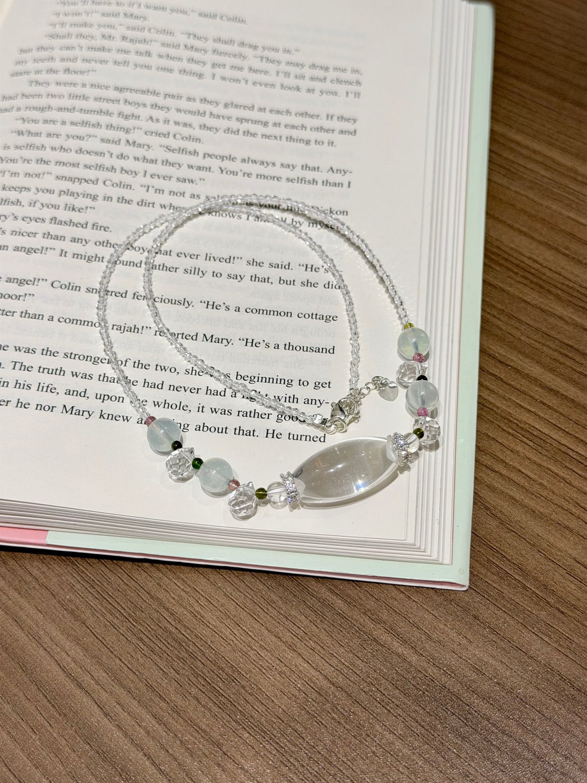 A silver-toned beaded necklace with a large teardrop-shaped clear stone and smaller pastel beads, displayed on an open book over a wooden surface.