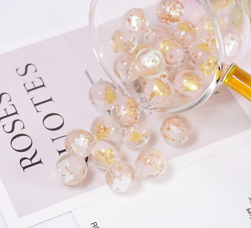 Clear round glass beads with embedded gold foil flakes, spilling from a clear glass jar onto printed paper.