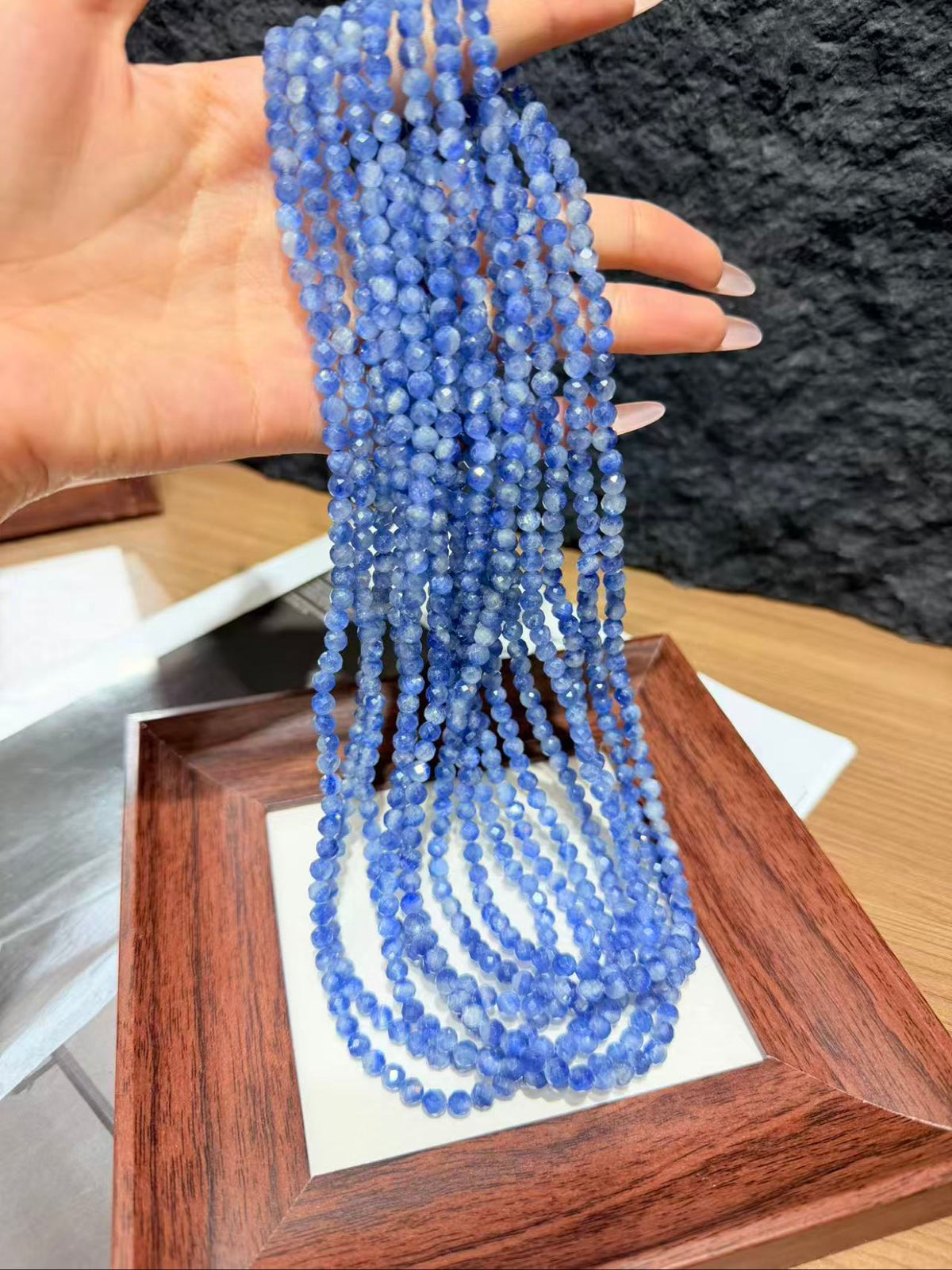 Hand holding long hanging strands of small blue faceted kyanite beads above a wooden display frame.