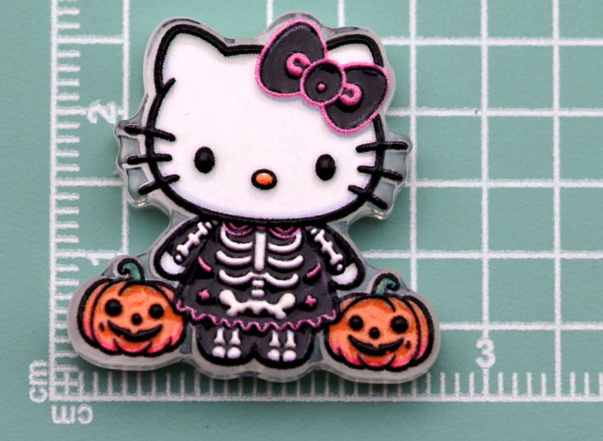 Hello Kitty skeleton acrylic charm with black and pink bow, skeleton outfit, and two pumpkins on green cutting mat