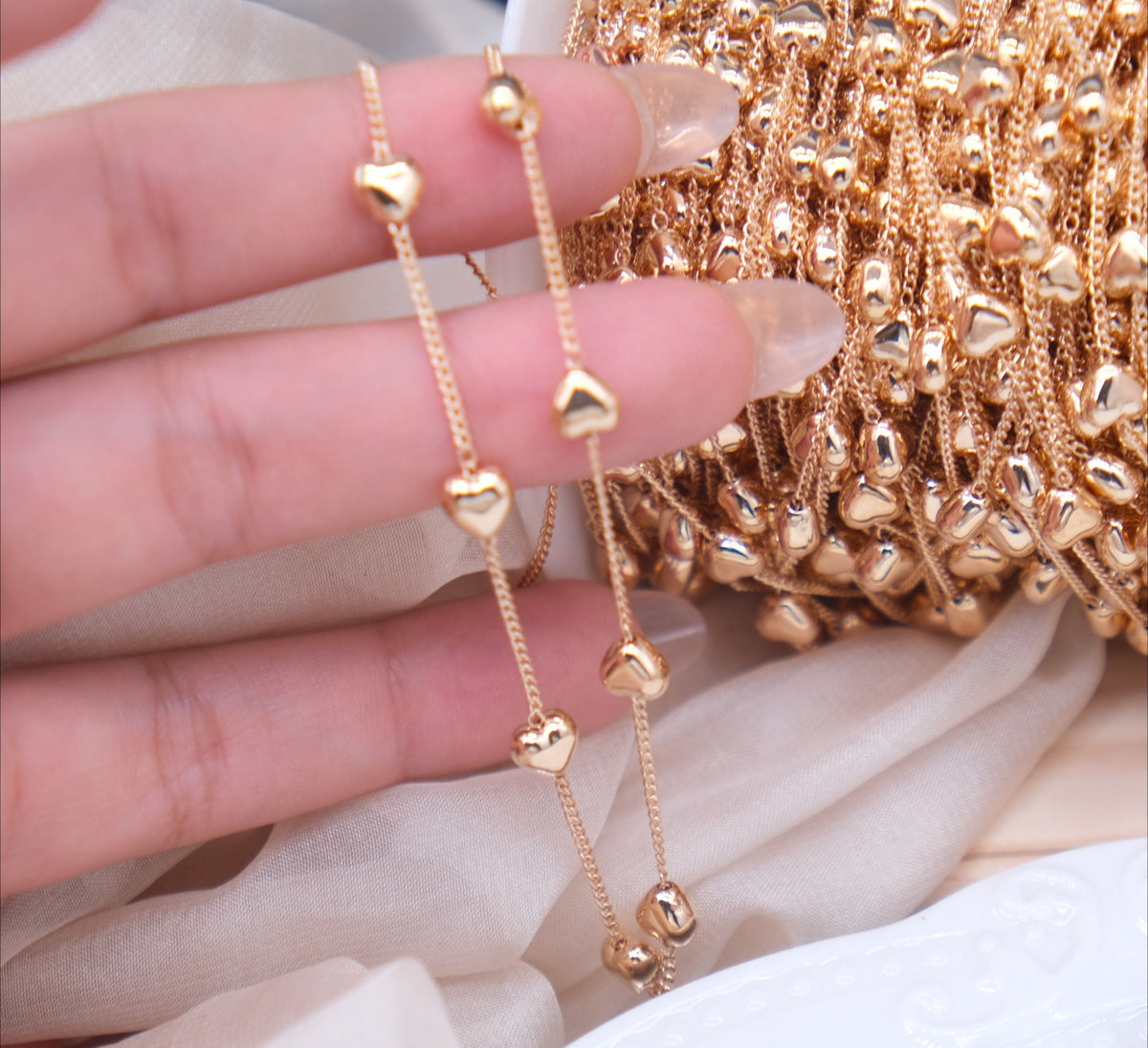 Close-up of fingers holding two rose gold heart-bead chains beside a spool of chain