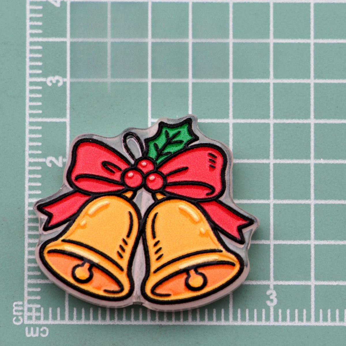 Christmas bell acrylic charm with two gold bells, red bow, and holly on green cutting mat