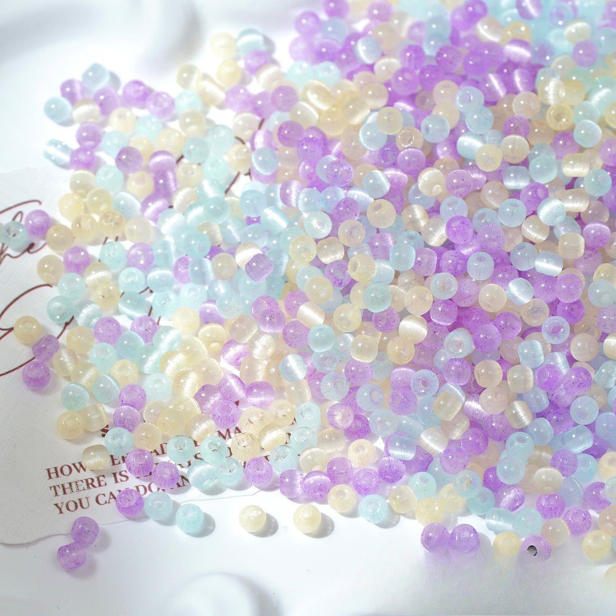 A pile of small translucent round beads in pastel lavender, light blue, and pale yellow, scattered on a white surface with a small piece of paper visible underneath.
