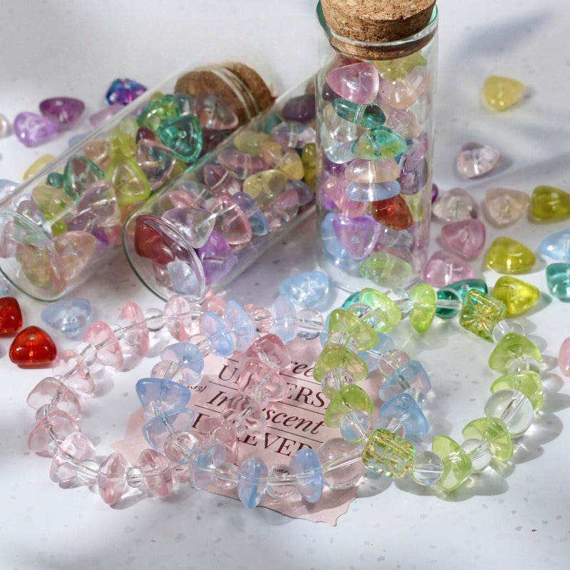 Colorful heart-shaped crystal beads shown as bracelets and small glass vials with cork stoppers on a light surface.