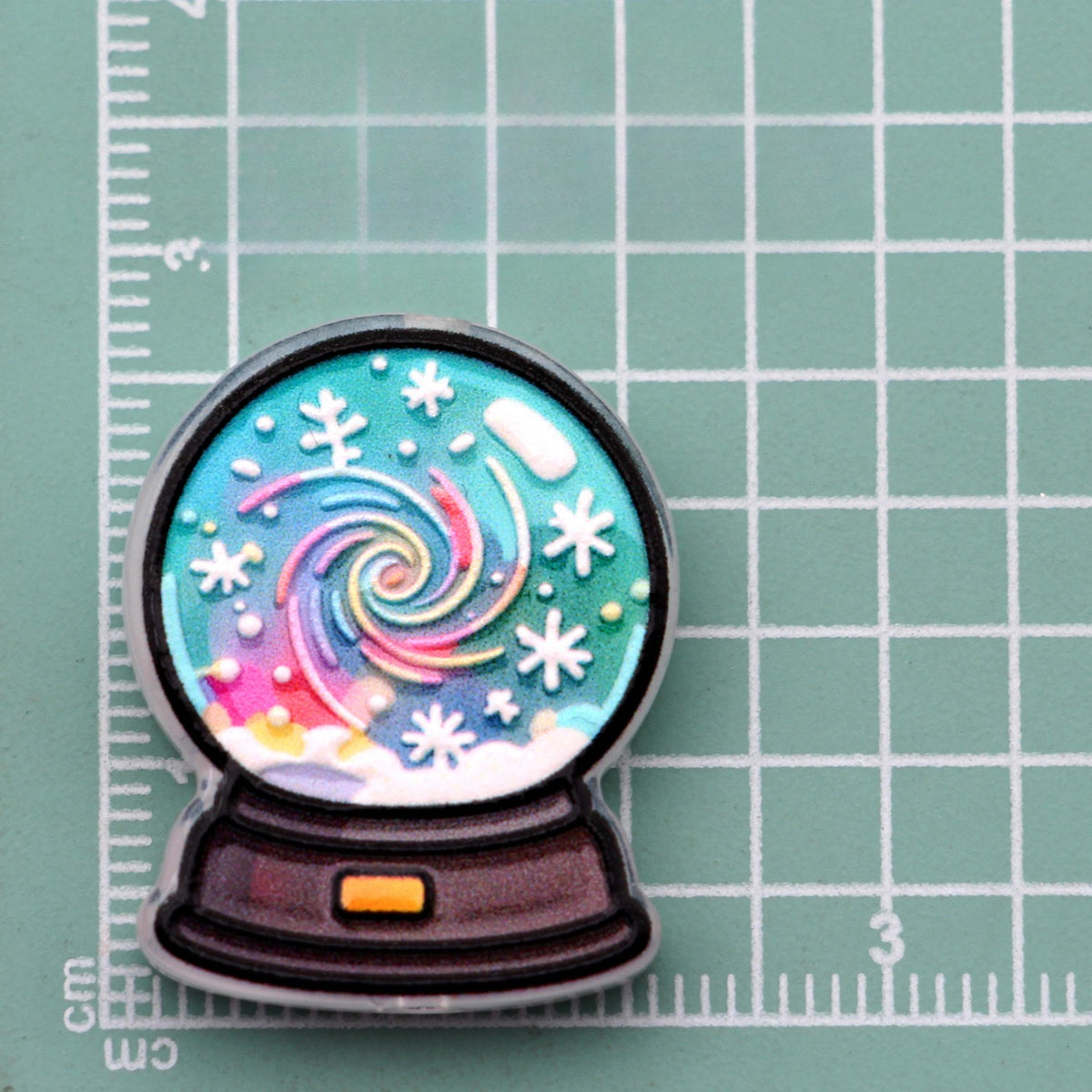 Snow globe acrylic charm with colorful spiral swirl and white snowflakes on green cutting mat