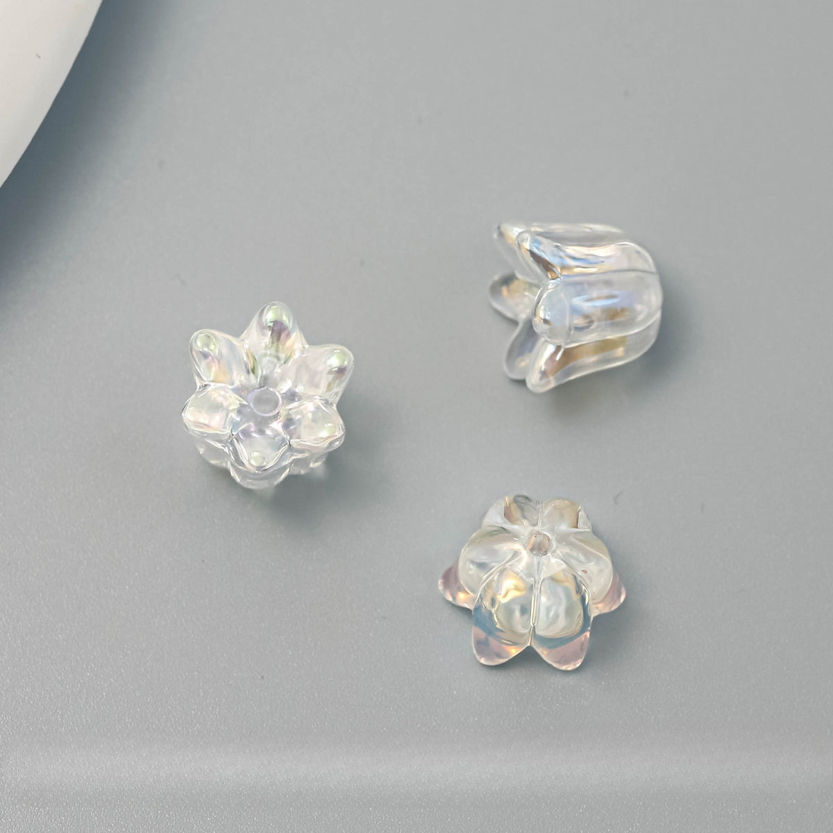 Three clear, iridescent flower-shaped craft beads arranged on a smooth gray surface.