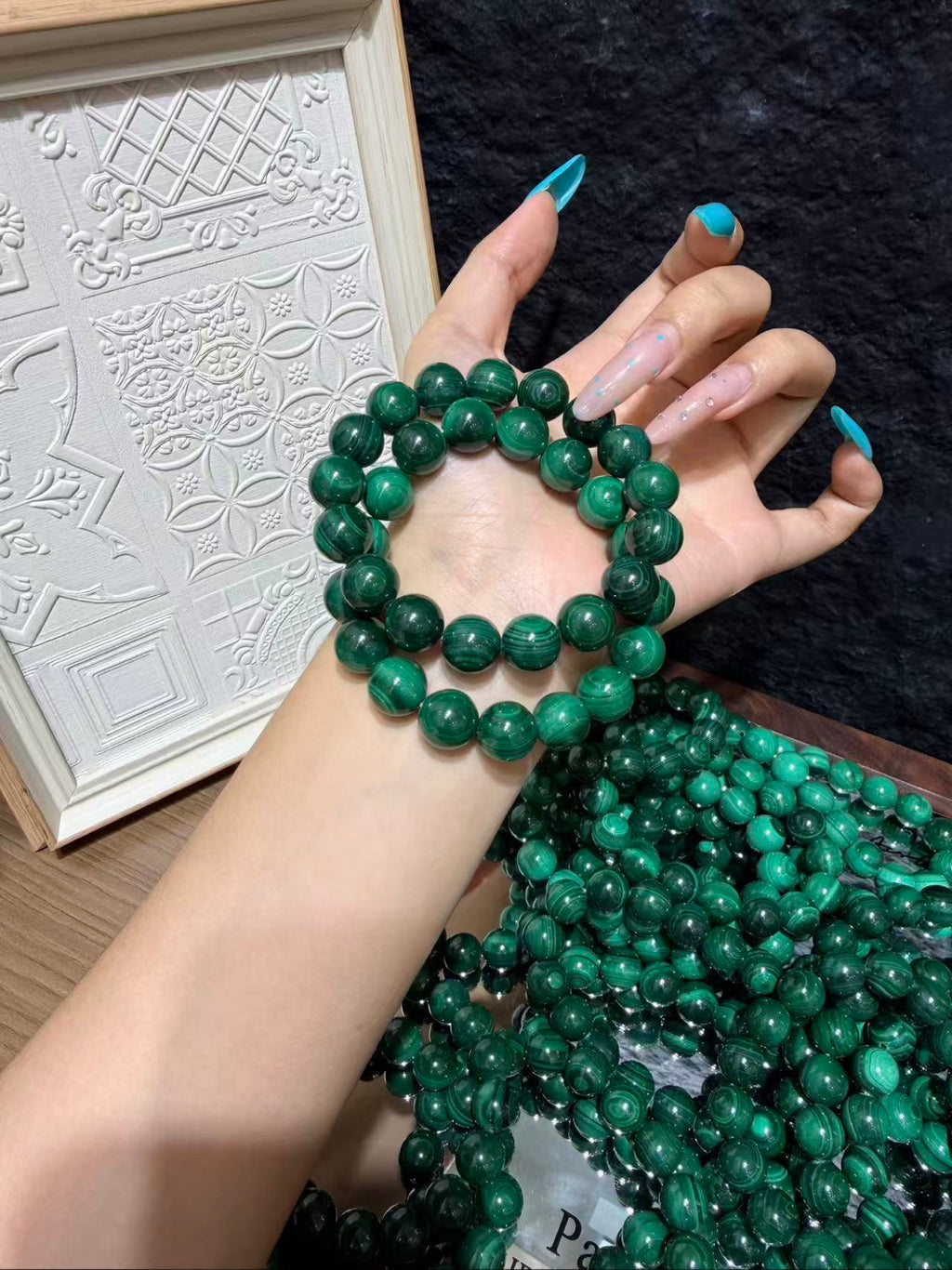 Hand with blue manicured nails wearing a double-strand bracelet of round green malachite beads with swirling patterns, held over a pile of matching loose malachite beads