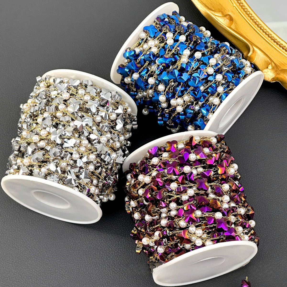 Three spools of butterfly chain in silver, royal blue, metallic purple with pearl accents on dark gray