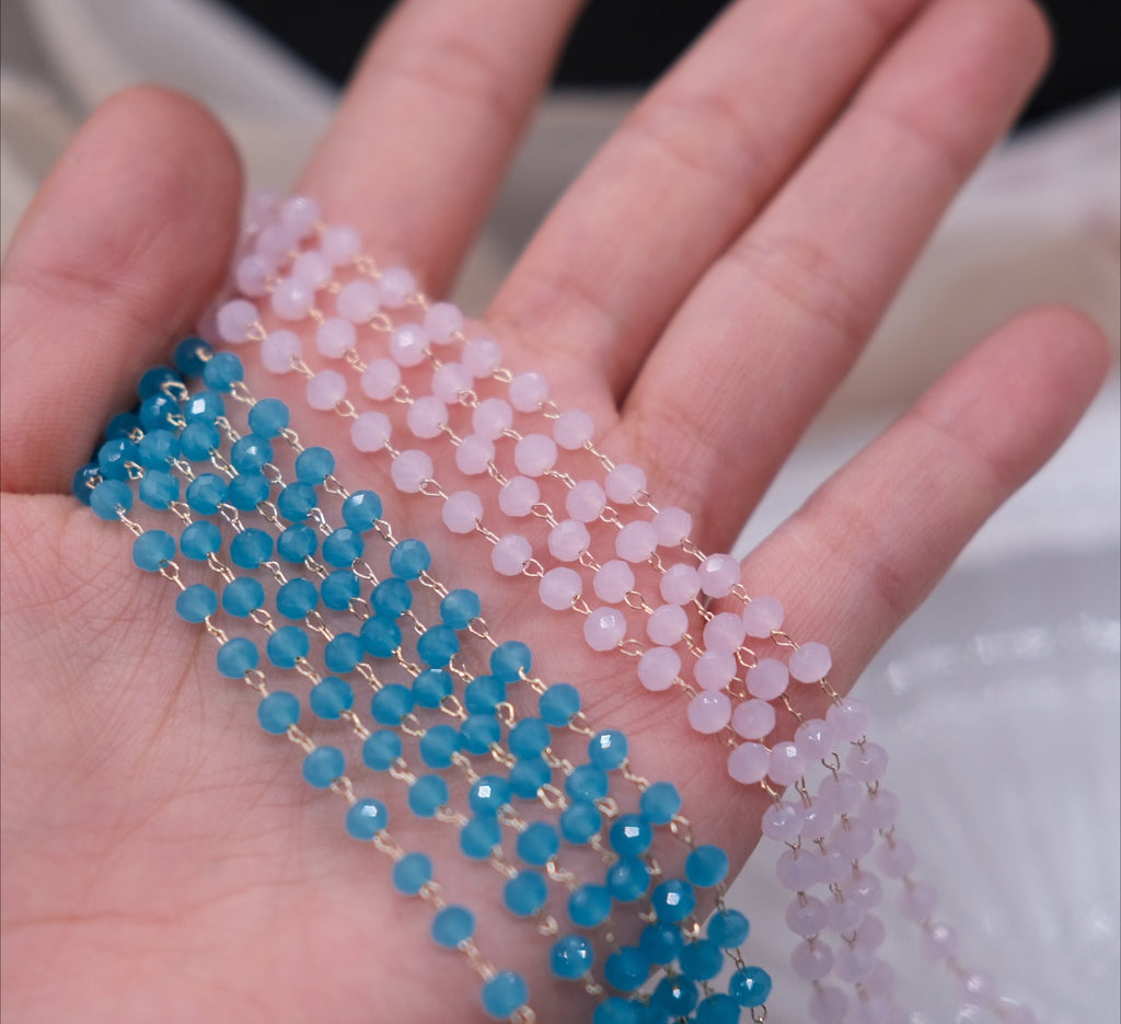 Two strands of faceted round beads draped across an open hand. One strand is light pink and the other is bright blue, both separated by thin gold-tone wire ties.