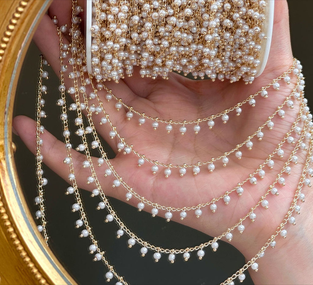 Hand holding gold 3mm white glass pearl chain strands draped over palm, gold mirror behind.