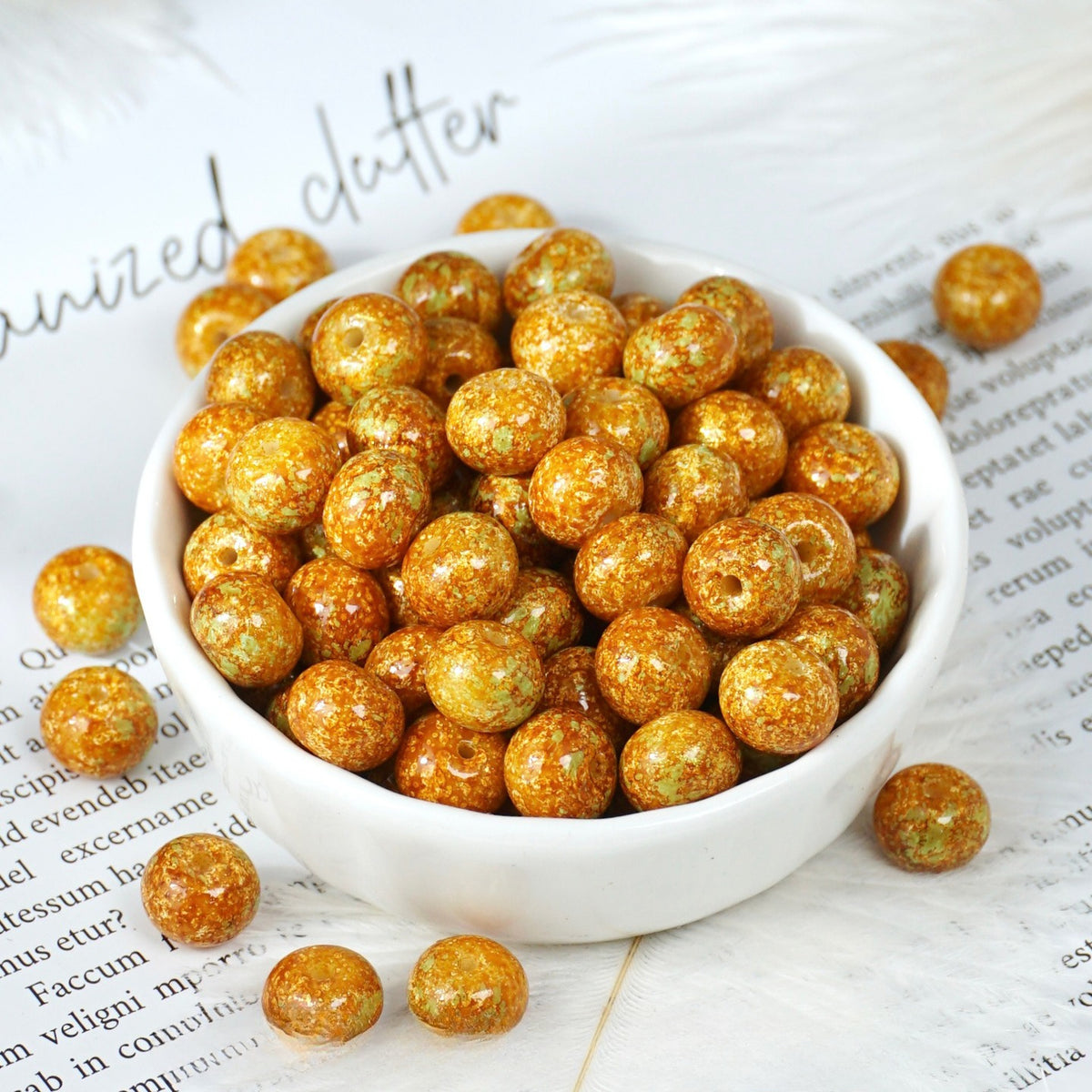 White porcelain bowl of small round golden-brown glazed beads, matching beads scattered on printed paper