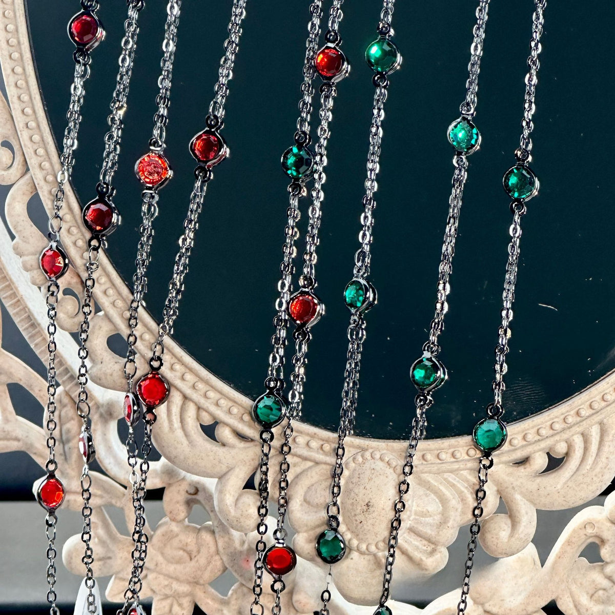 A close-up of a beaded curtain necklace with multiple vertical strands of silver-toned chains, alternating red and green faceted beads, draped over an ornate cream-colored mirror frame.