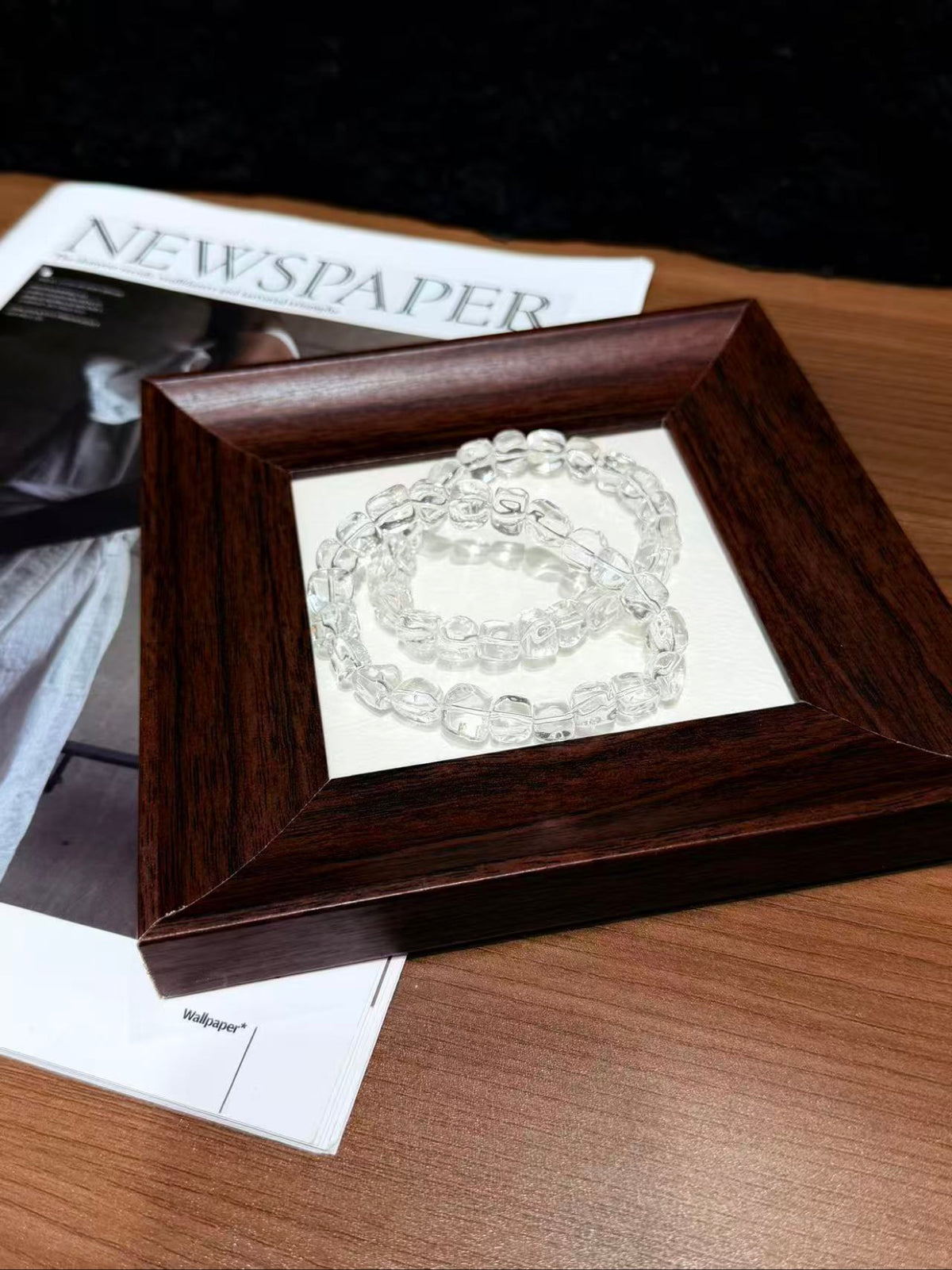 White quartz jelly-bean nugget bracelets coiled and displayed in a dark wooden frame on paper, beside a newspaper on a wooden table