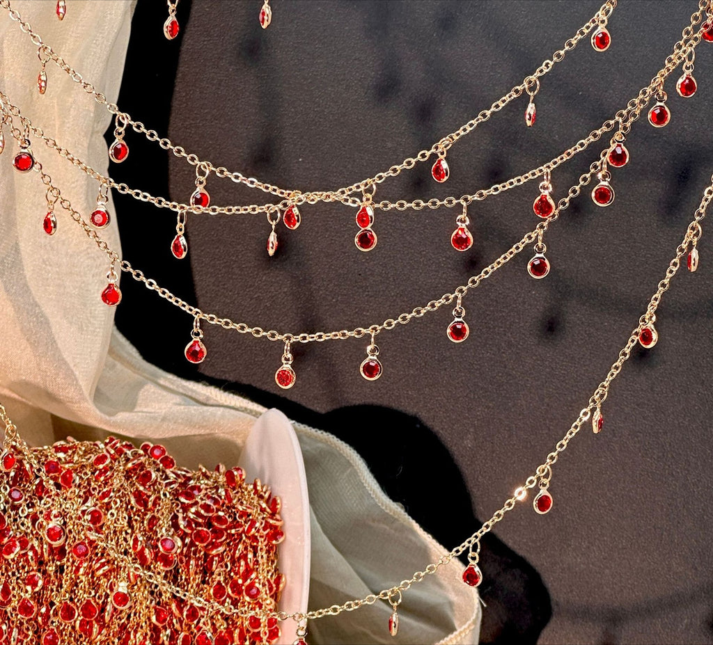 Gold-toned chain garlands with multiple dangling red teardrop-shaped stones, draped across a dark surface with sheer white fabric and a matching embellished textile partially visible.