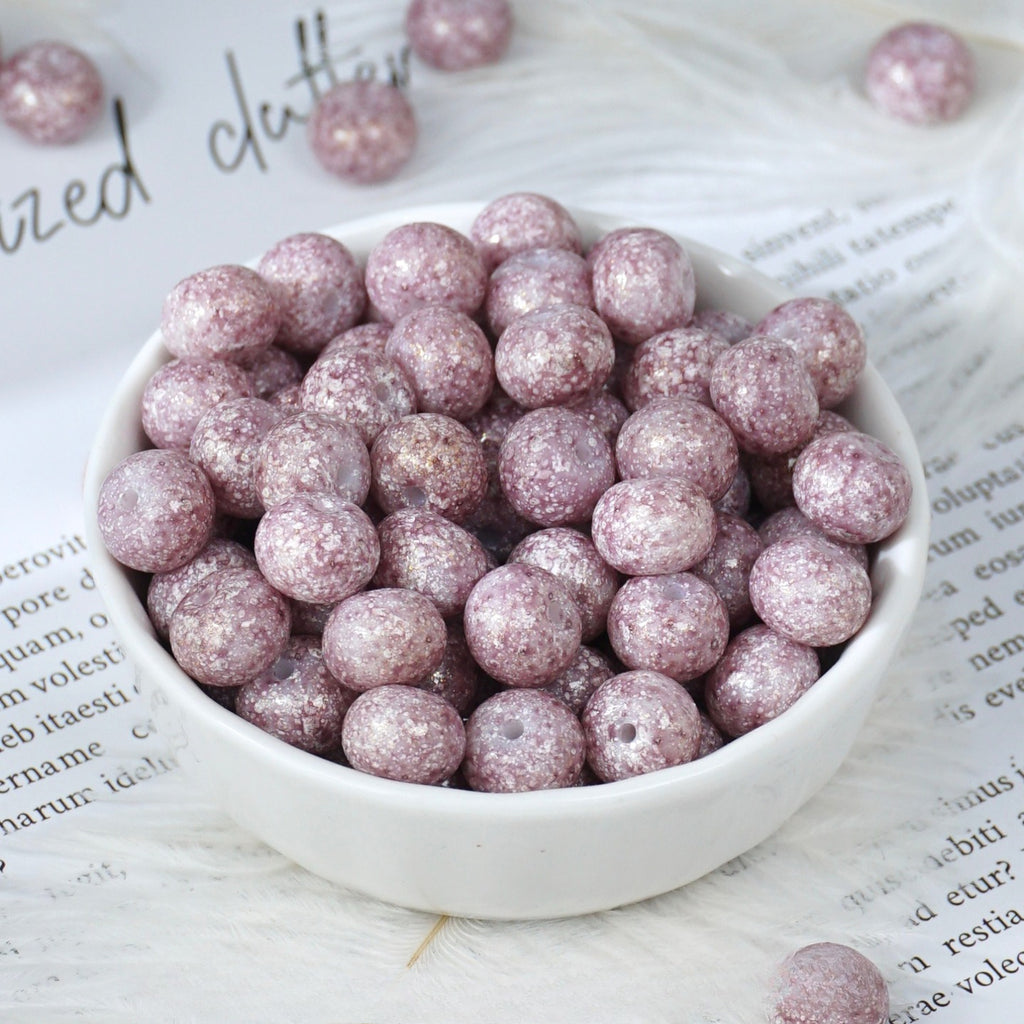 White porcelain bowl filled with round glittery lavender-purple glazed porcelain beads, a few loose beads scattered nearby