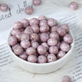 White porcelain bowl filled with round glittery lavender-purple glazed porcelain beads, a few loose beads scattered nearby