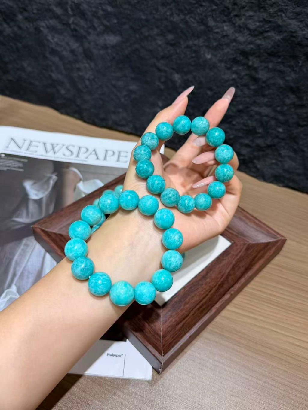 Hand displaying two turquoise amazonite round-bead bracelets, one worn on the wrist, over a wooden tray and newspaper