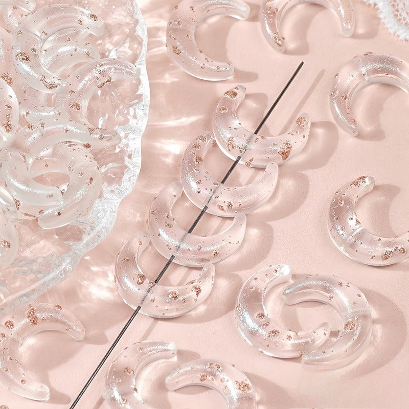 Clear glazed glass crescent moon beads with gold glitter, some strung on a wire and others on a pink surface with a clear dish