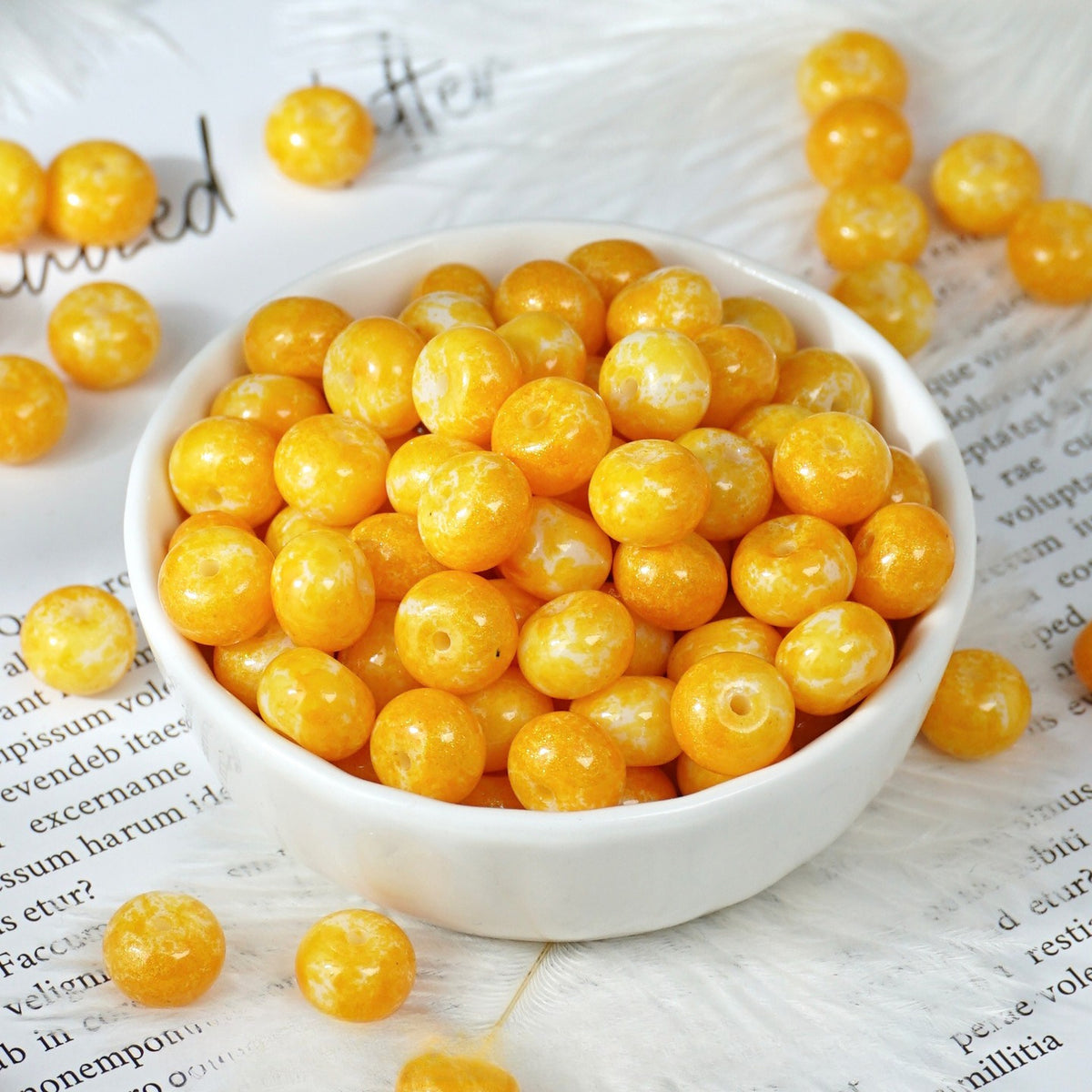 Bright yellow-orange round beads with white marbling in a small white porcelain bowl, some scattered on printed paper