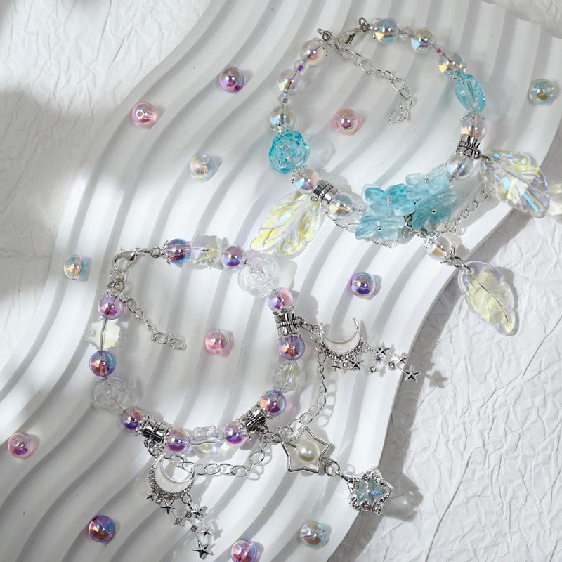 Three beaded charm bracelets arranged on a white wavy surface. The bracelets feature silver-tone chains with iridescent beads, translucent blue flower charms, clear leaf-shaped beads, and celestial charms including a moon, star, and pearl.