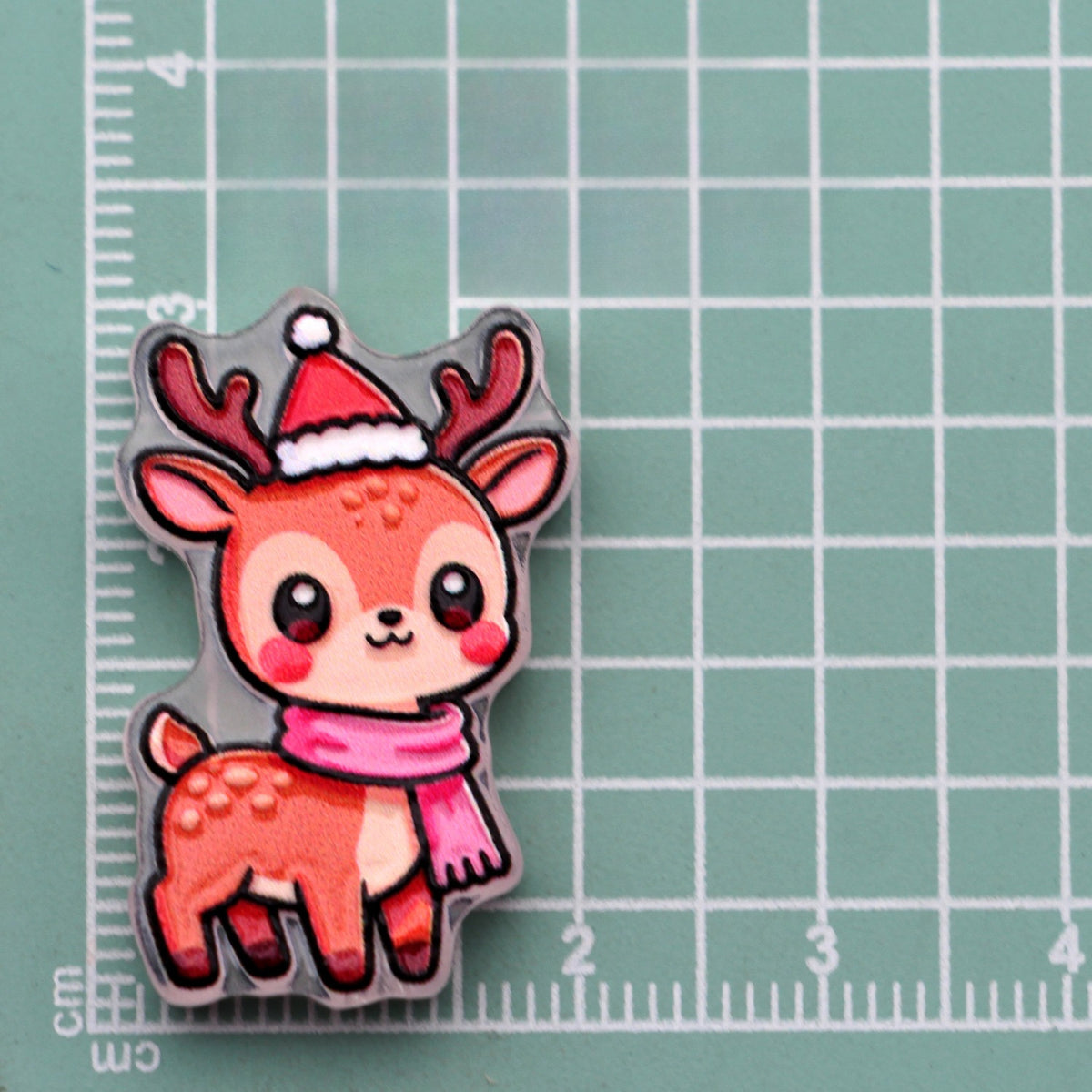 Baby reindeer acrylic charm with red Santa hat and pink scarf on green cutting mat