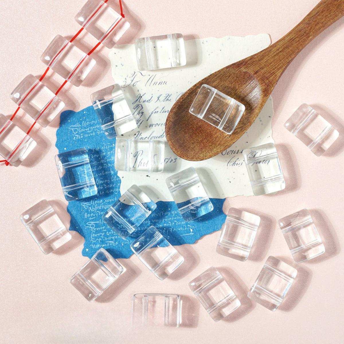 Clear square glass beads on pink with a wooden spoon, blue and white paper and red string